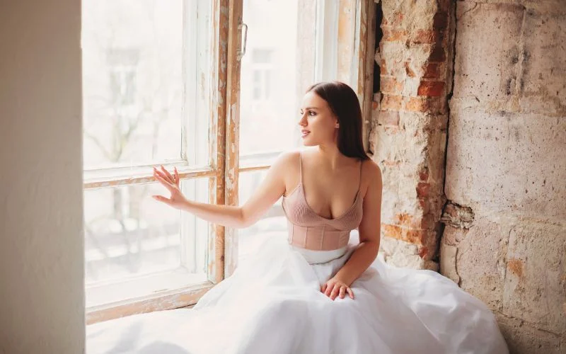 Nude bridal photo of a bride in a flowing gown seated by a large window