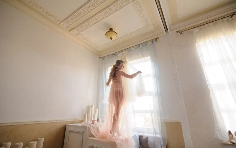 Nude bridal pictures of a bride in a flowing white robe standing by a sunlit window