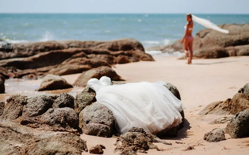 Nude wedding pics of a bride's wedding dress laid on coastal rocks with the bride standing at the shoreline