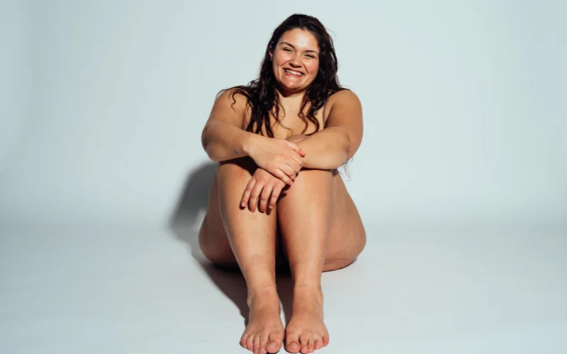 Curvy nude boudoir pose with woman seated gracefully on a bed, soft window light highlighting her silhouette
