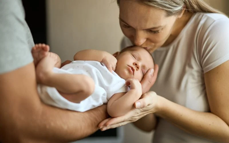 Newborn poses with parents showing a mother cradling and kissing her baby's forehead