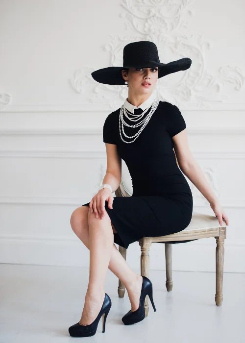 Woman in a black dress and wide-brim hat with pearl necklaces, seated on a stool