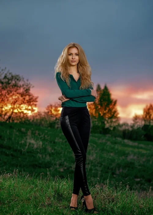 Woman in a green top and black pants standing in a field at sunset, color-coordinated with the sky