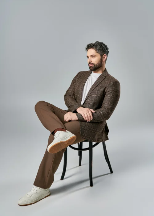Man in a brown suit seated on a stool, lit with soft studio lighting