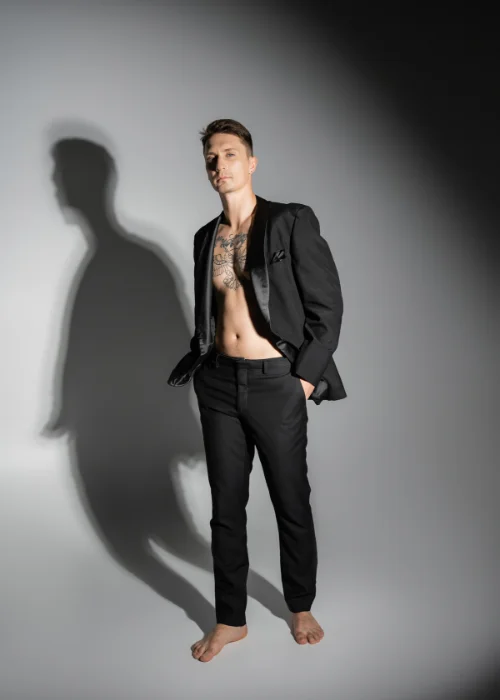 Man in an open dark jacket casting a harsh shadow against a studio wall