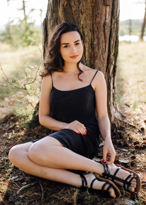 Woman sitting against a tree trunk in a black dress, hands resting naturally in her lap