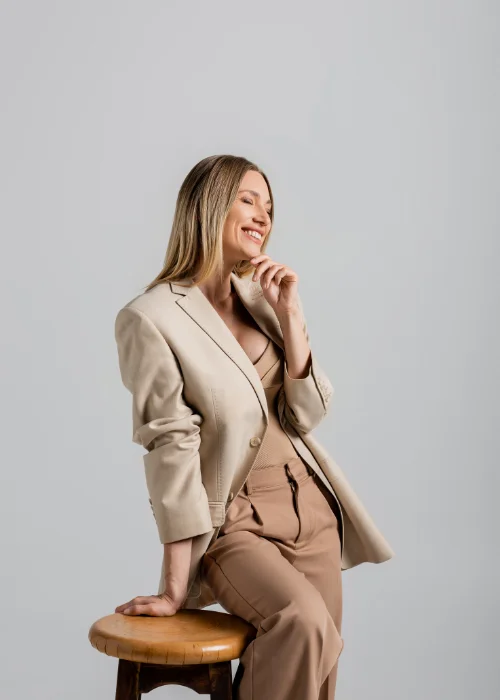 Full body portrait of a woman in a beige suit seated on a stool, shown in side profile