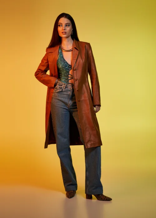 Full length portrait of a woman in a brown leather coat standing tall against a yellow backdrop