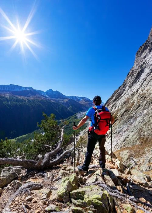Hiker standing on a rocky mountain trail with a backpack while admiring panoramic views under a bright blue sky.