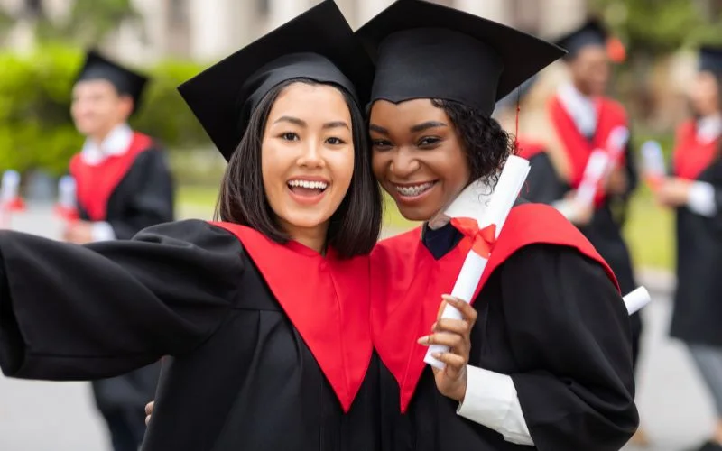 Two graduates in caps and gowns holding diplomas close together, smiling at the camera