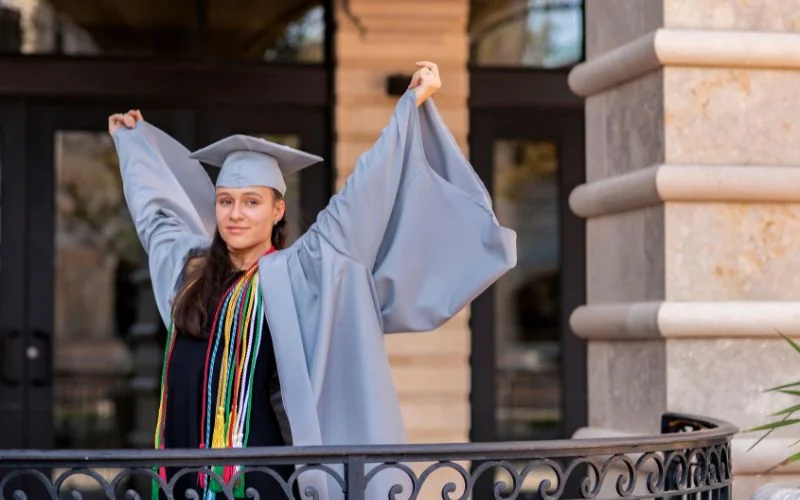 Graduation photo poses male style with a graduate holding her gown open, standing against a stone railing