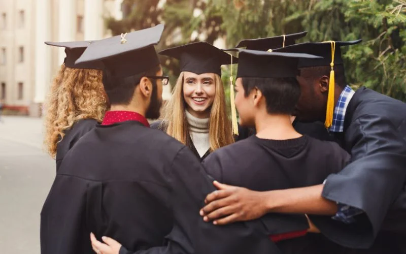 Group of graduates embracing in a circle hug wearing caps and gowns on campus
