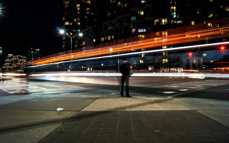 City street at night with a person standing still while car light trails blur past on a long exposure