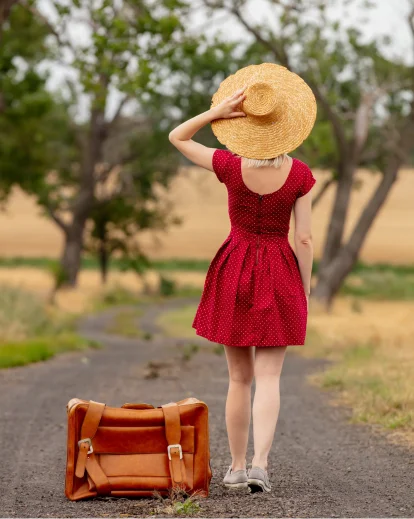 Woman in a red dress and wide-brim hat standing on a road beside a vintage suitcase