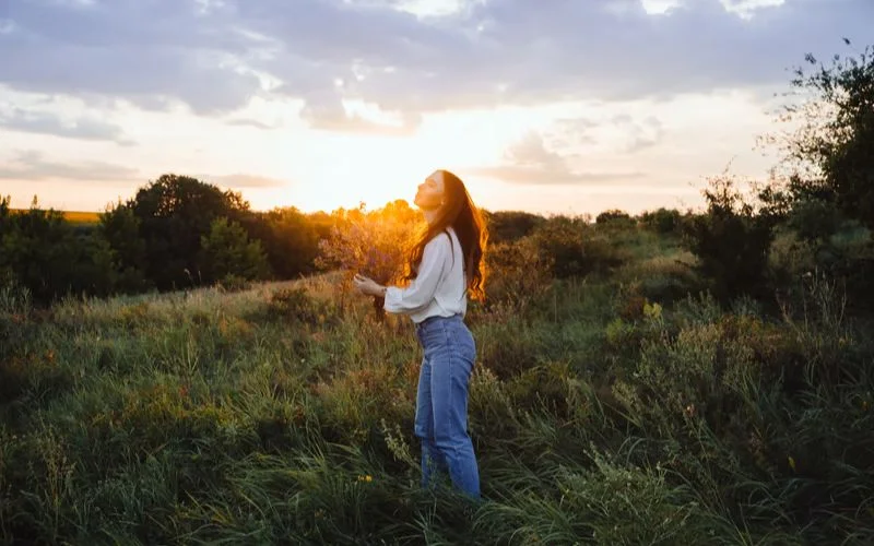 Woman in a white top standing in a field, shielding her eyes from bright sunset light with visible highlight and shadow contrast