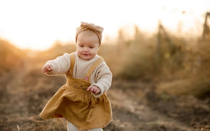 Toddler walking through a sunlit meadow with soft, warm golden hour light behind them