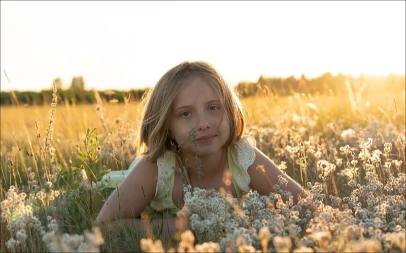 Close-up portrait of a girl lying in a flower field with soft, shadow-free golden hour lighting on her face