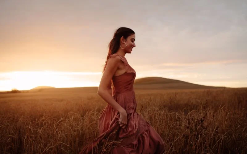 Woman in a red dress walking through a golden field with a visible sun lens flare across the frame