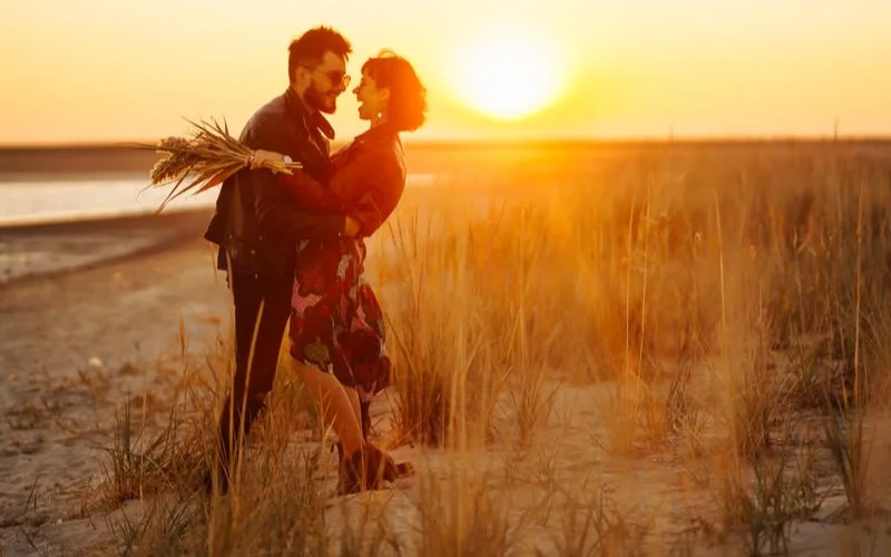Couple embracing on a beach at sunset holding tall grass, with the sun positioned directly behind them